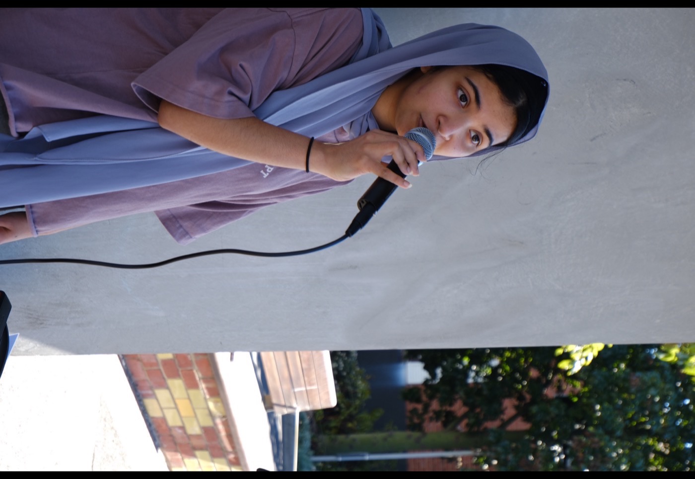 Omari singing into a microphone against a rendered wall, outdoors in daylight.